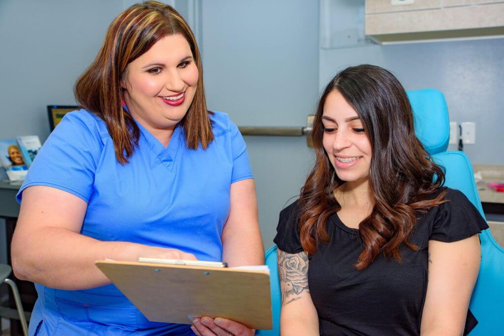 Route 32 Dental team member wearing blue office uniform pointing to a clip board in her hand in a consultation with a patient wearing a black top who is sitting in the dental treatment chair