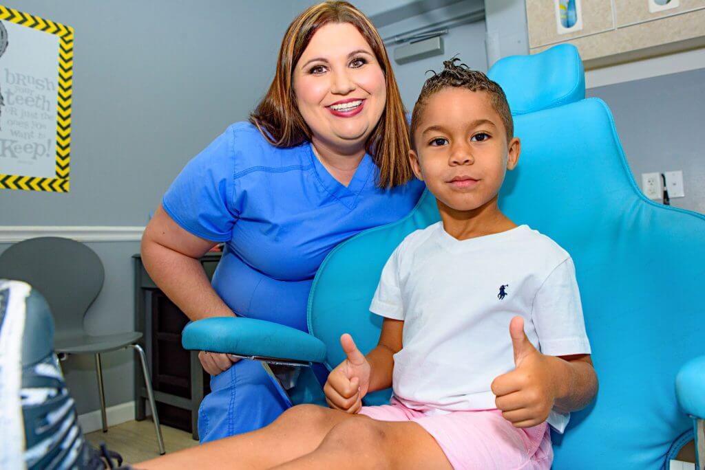 A smiling Route 32 Dental team member wearing blue uniform sitting next to a young patient who is sitting in the blue dentist treatment chair and signaling holding both his thumbs up