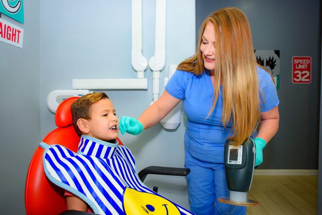 Route 32 Dental team member in blue uniform and green exam gloves holding an X-Ray machine in one hand, while positioning the X-Ray film with the other hand  in the mouth of a young patient who is wearing the protective X-Ray apron with blue stripes and a smiley face print, and is sitting on a red dentist chair