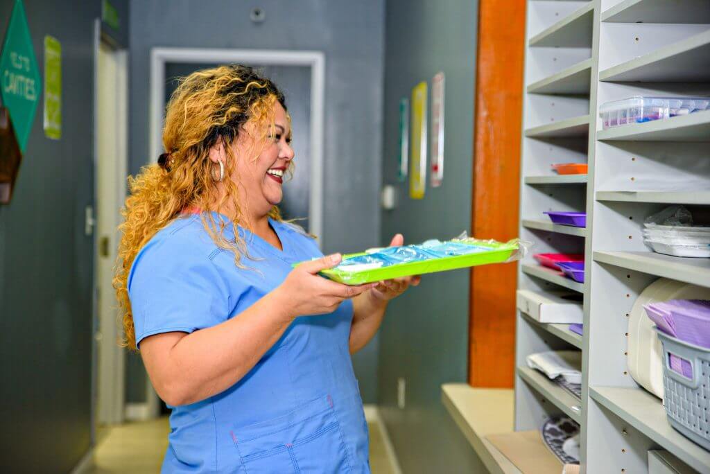 Route 32 Dental team member smiling while holding a lime green color tray with invisalign on, standing in front of the shelving cabinet that holds many colorful assortment of additional dentist exam and treatment tools set ready for use in the patient room